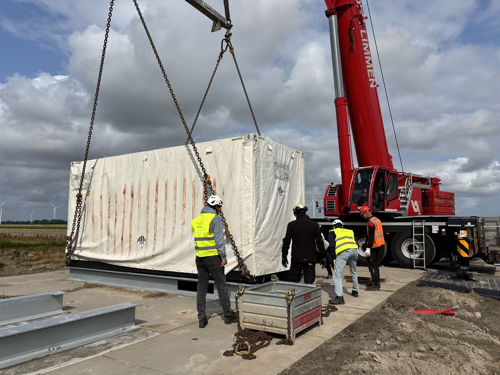 Monteurs met helm en veiligheidshesje begeleiden een batterijcontainer die met een kraan op de fundering zakt