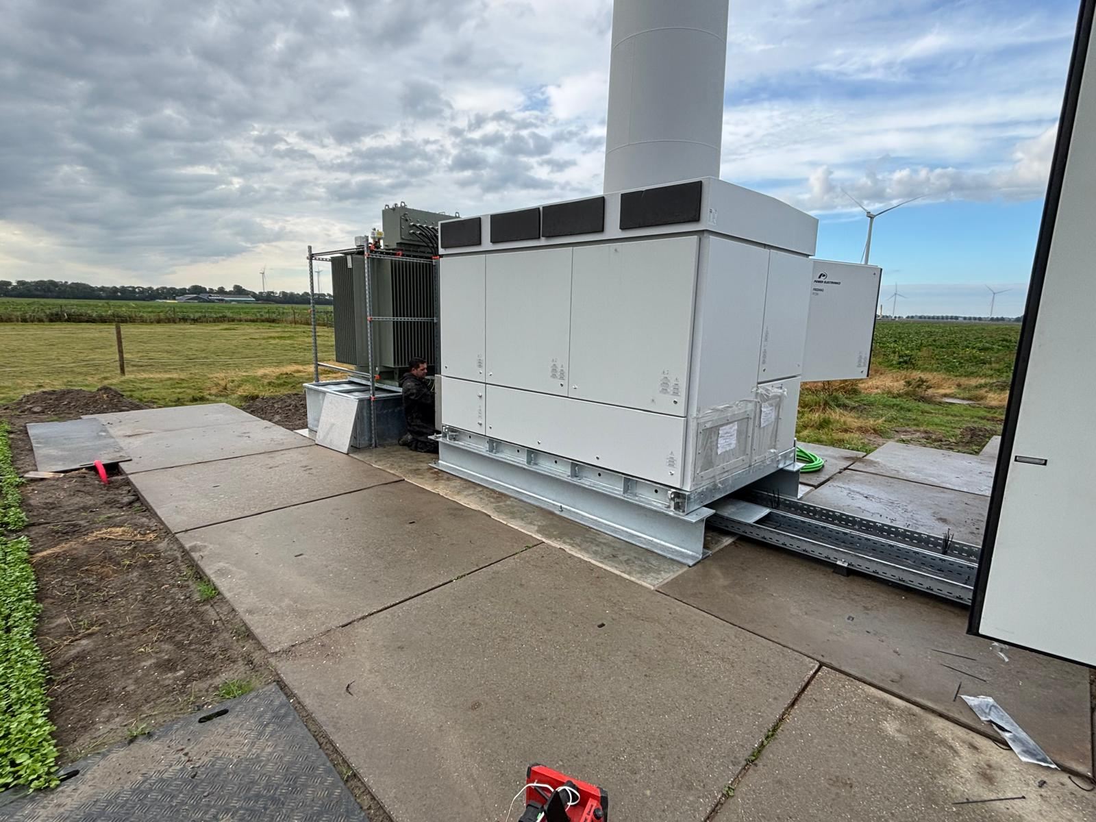 Batterijcontainers en trafo binnen een hekwerk, naast een windturbine